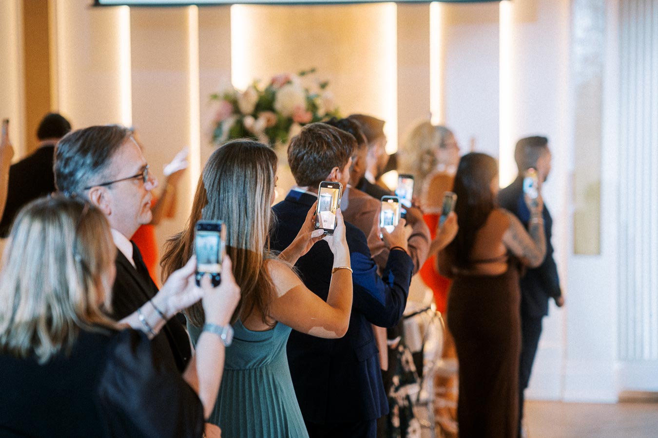 Guests at a wedding ceremony using smartphones to capture photos, standing in formal attire inside a beautifully decorated