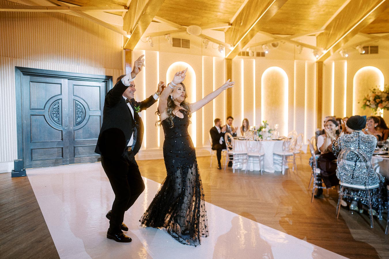 Elegant couple joyfully dancing at a stylish indoor celebration, surrounded by wedding guests seated at beautifully