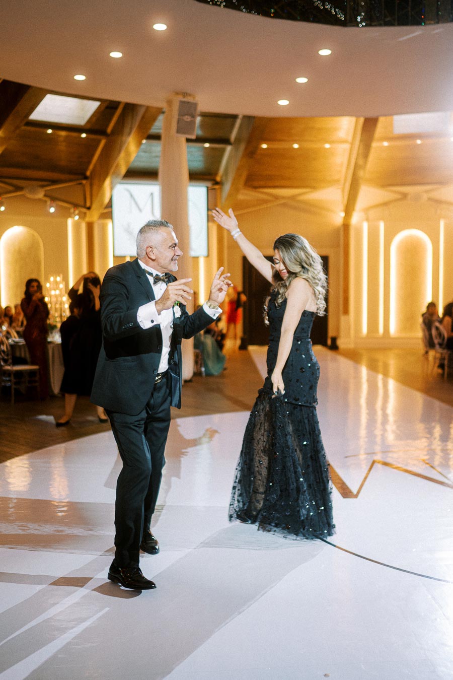 A well-dressed man and woman joyfully dance in a beautifully lit ballroom, surrounded by elegantly dressed guests at a