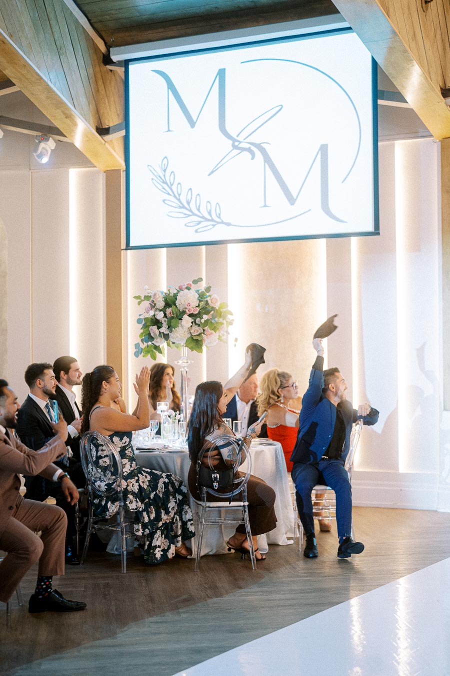 Wedding guests cheer and raise hands seated around a dining table, dressed in formal attire, with festive decor and a