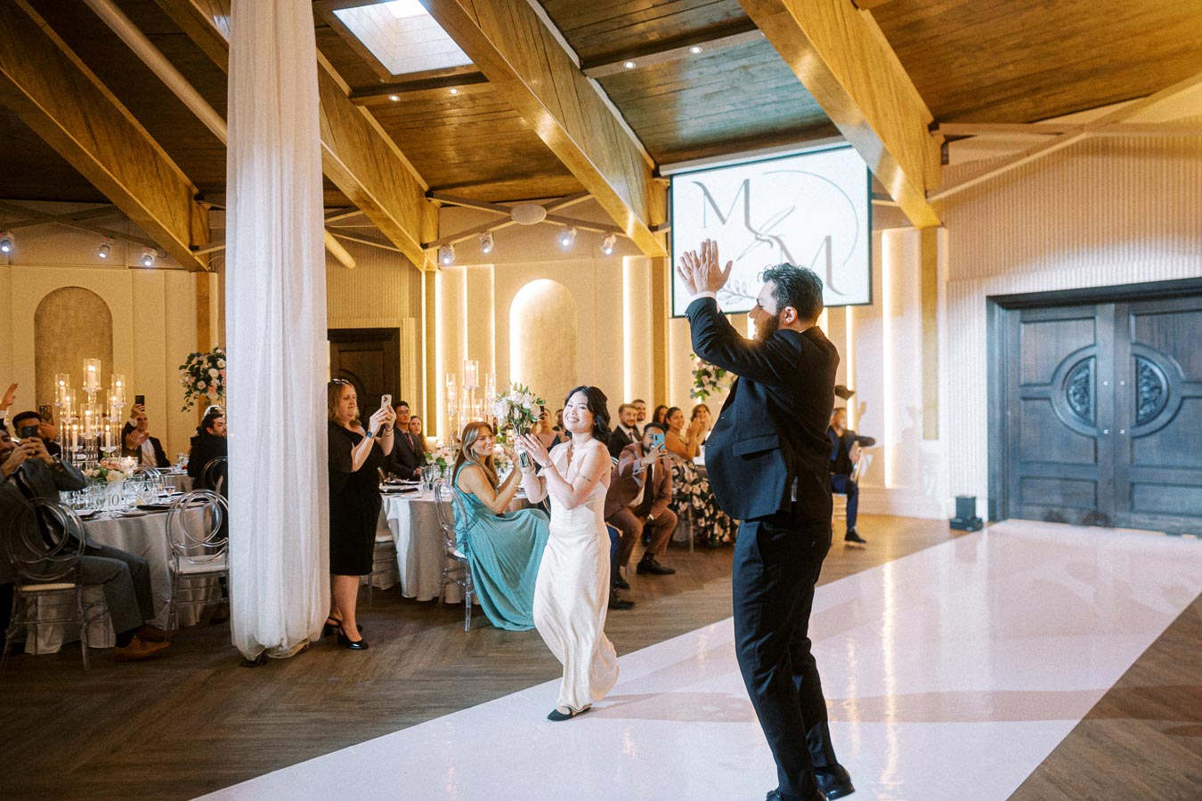 Wedding reception dance celebration in a beautifully lit venue with happy guests clapping and capturing the moment.