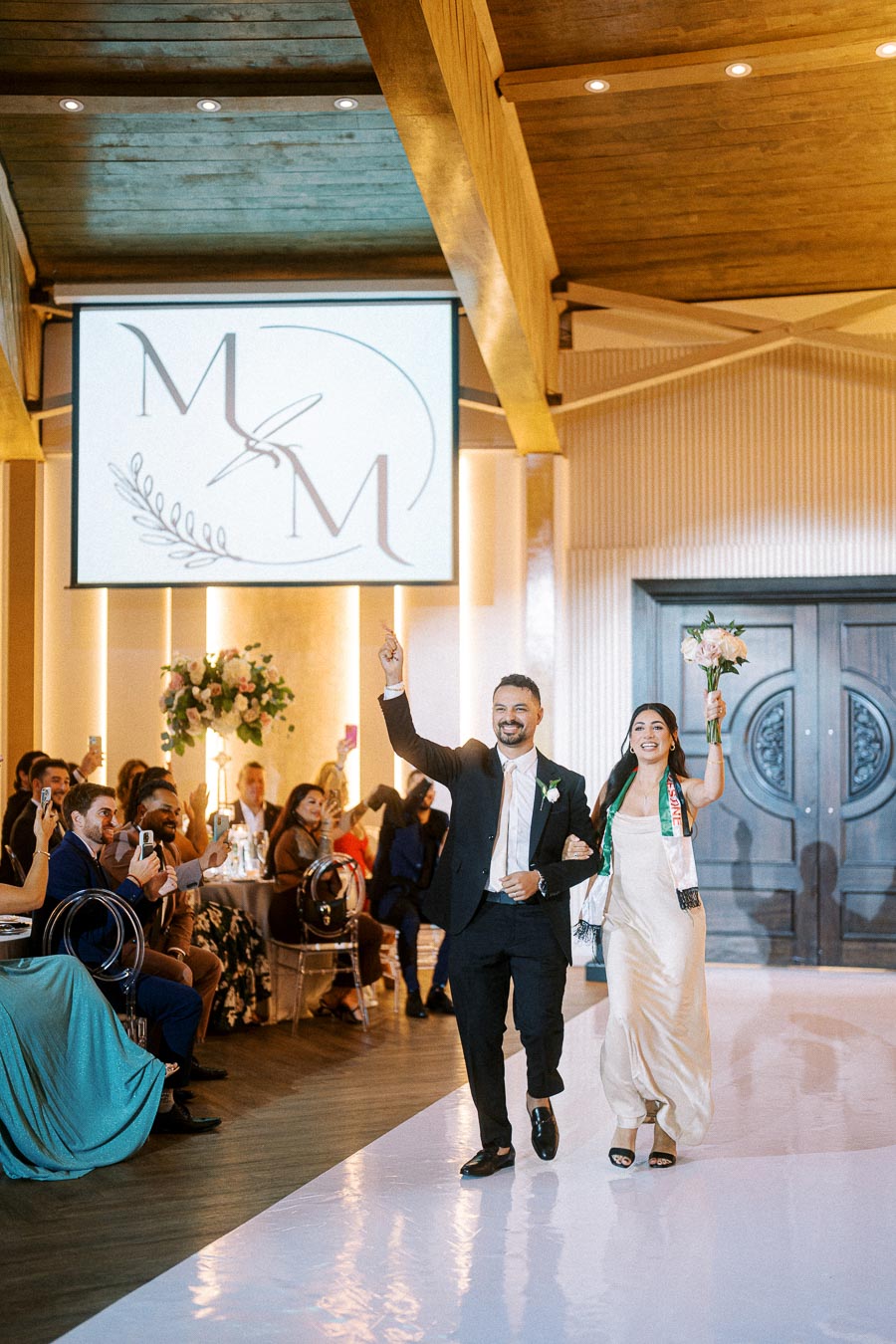 Couple celebrating their wedding entrance with joyful expressions, surrounded by cheering guests in elegant attire, under a
