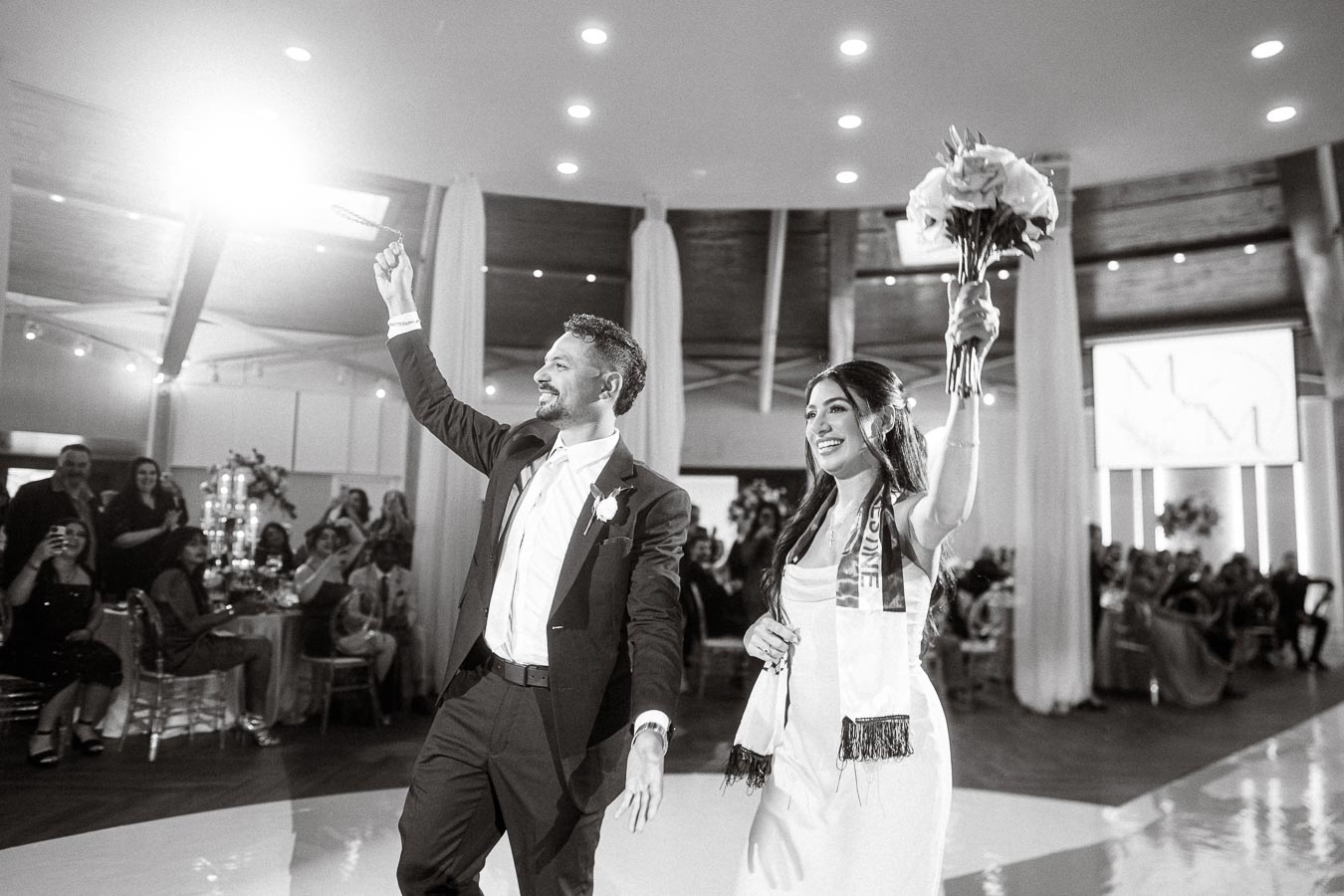 A joyful couple enters a wedding reception, smiling and waving at guests. The groom wears a suit and the bride holds a