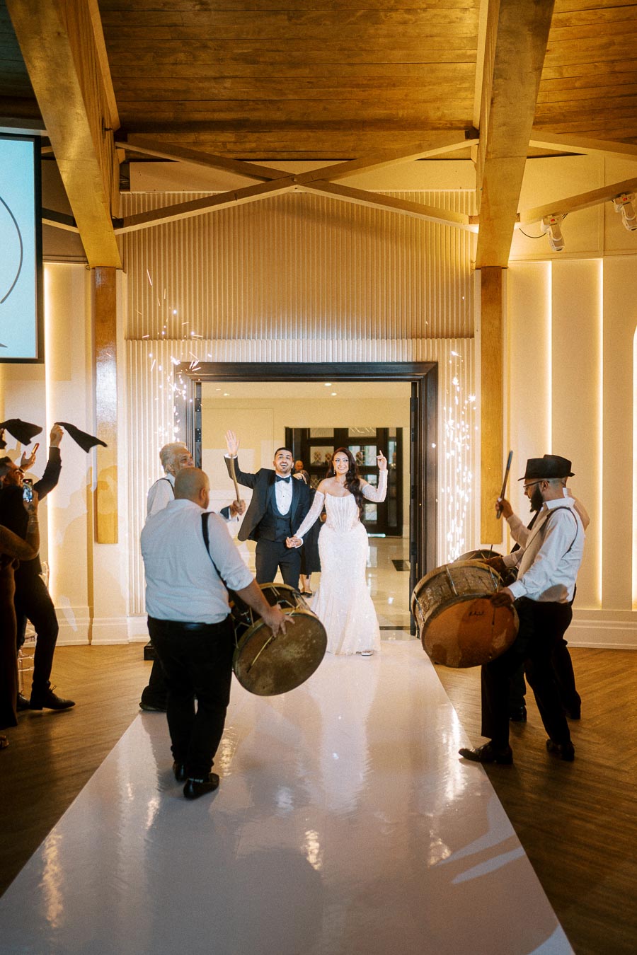A bride and groom jubilantly enter a wedding reception under a canopy of sparklers, accompanied by musicians playing drums,