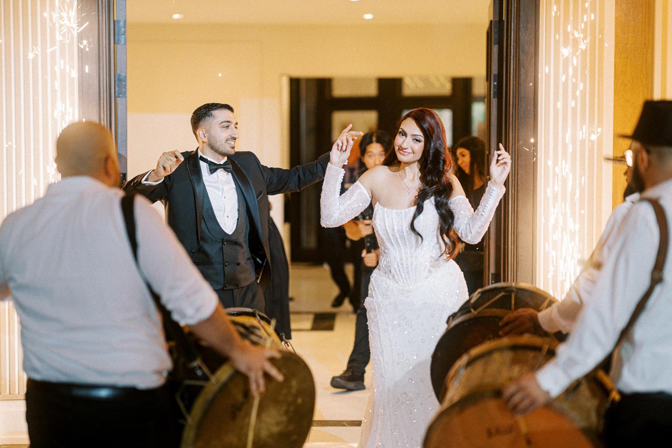 Bride and groom joyfully entering their wedding reception with musicians playing drums, surrounded by sparkler decorations.