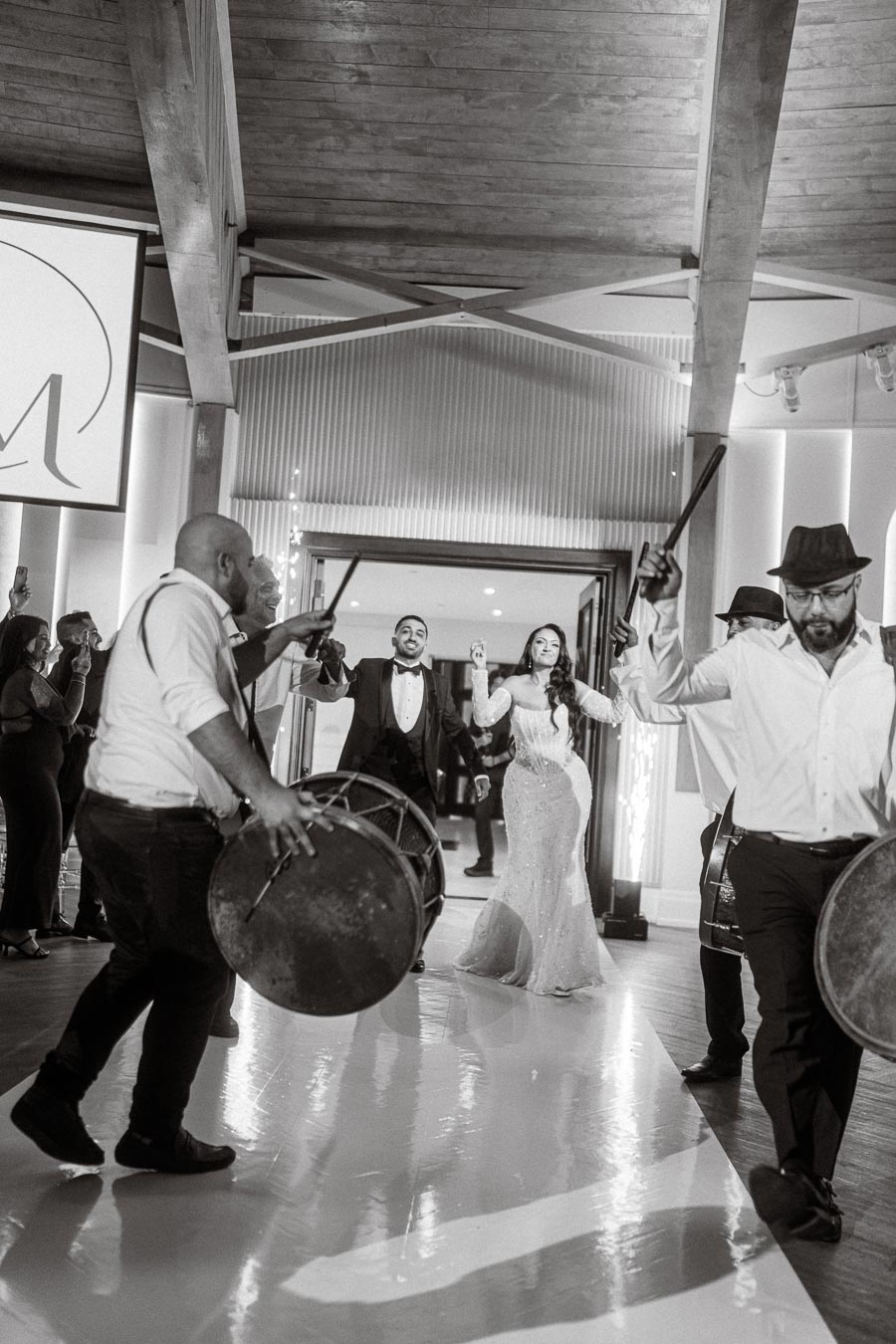 A bride and groom joyously dance into their wedding reception, accompanied by drummers in a lively celebration. The elegant