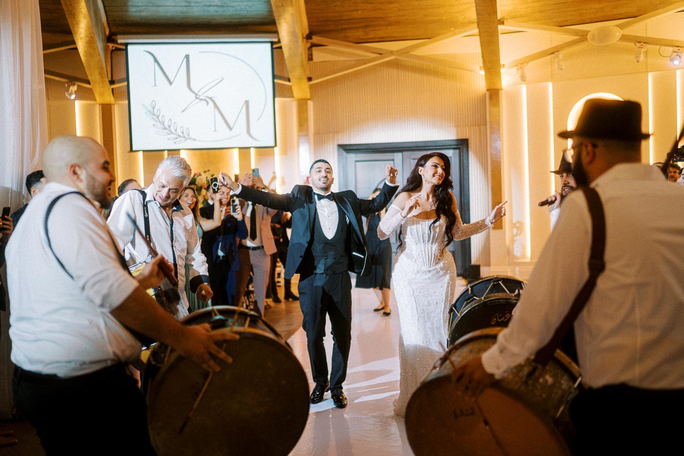 A joyful bride and groom dance surrounded by guests and drummers at a lively wedding reception, with a monogrammed logo