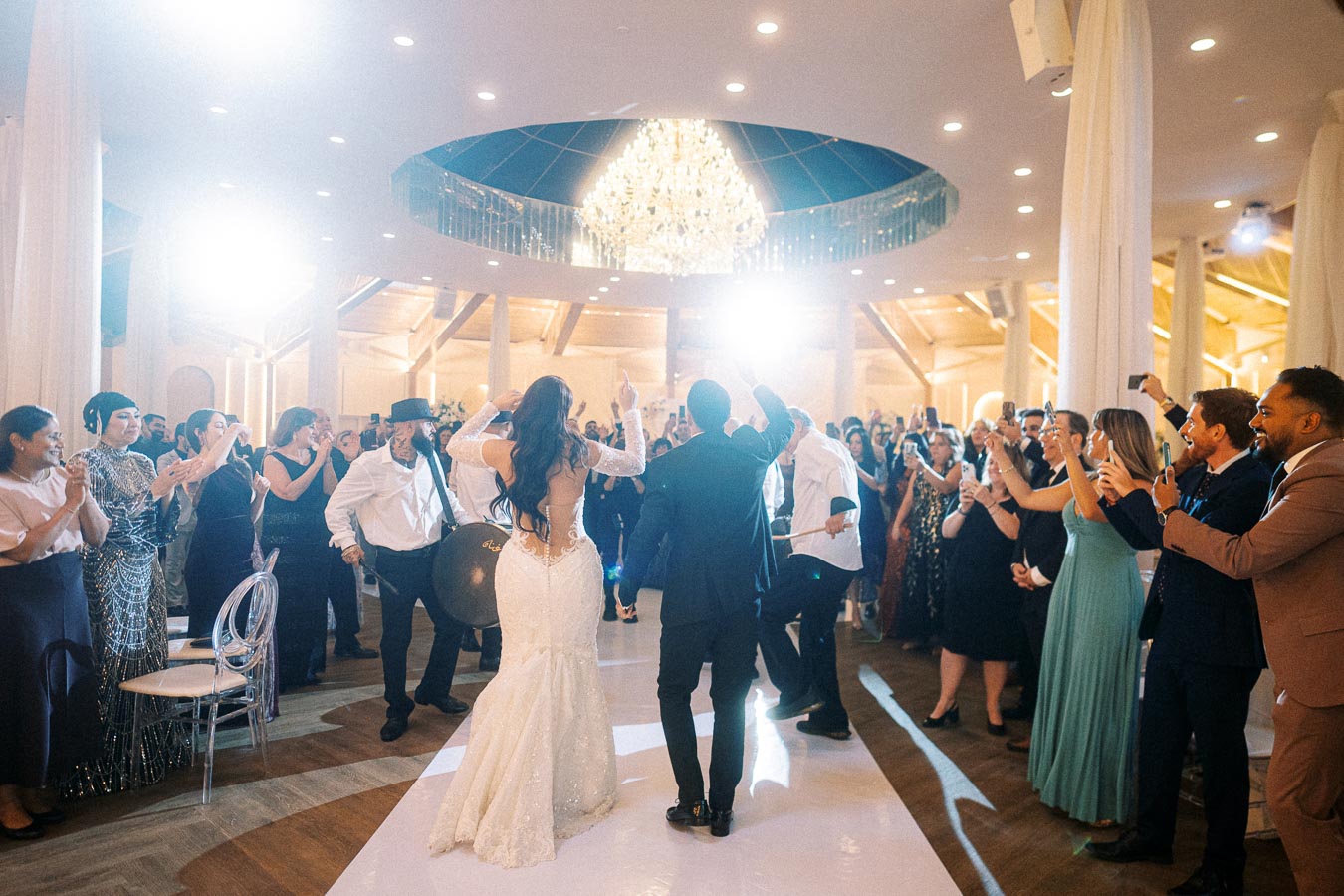 A couple dancing at a wedding reception surrounded by guests, with everyone clapping and taking photos in a beautifully