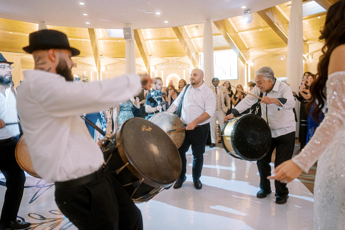 A lively indoor event with musicians playing traditional drums and guests in formal attire dancing, set in a bright venue