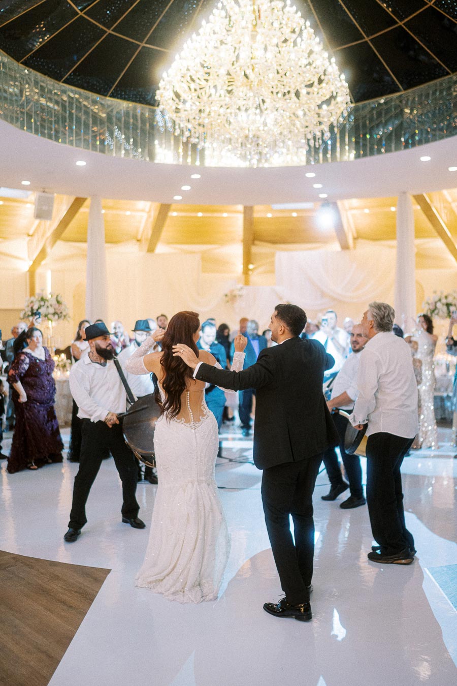 A bride and groom joyfully dancing at their wedding reception under a grand chandelier, surrounded by guests and musicians