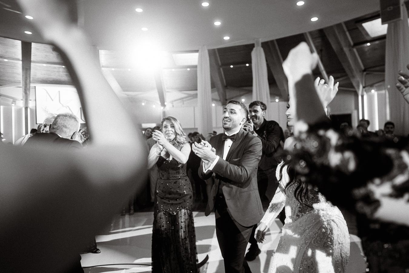 A joyful group of people dressed in formal attire, clapping and celebrating at a lively indoor event or wedding reception,