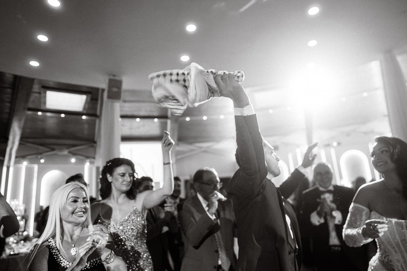 Black and white photo of a lively wedding celebration. Guests are joyfully dancing under bright lights. One person holds a