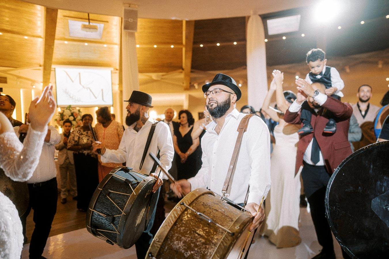 Two drummers perform energetically at a lively indoor event, entertaining a diverse crowd of joyful guests, including a