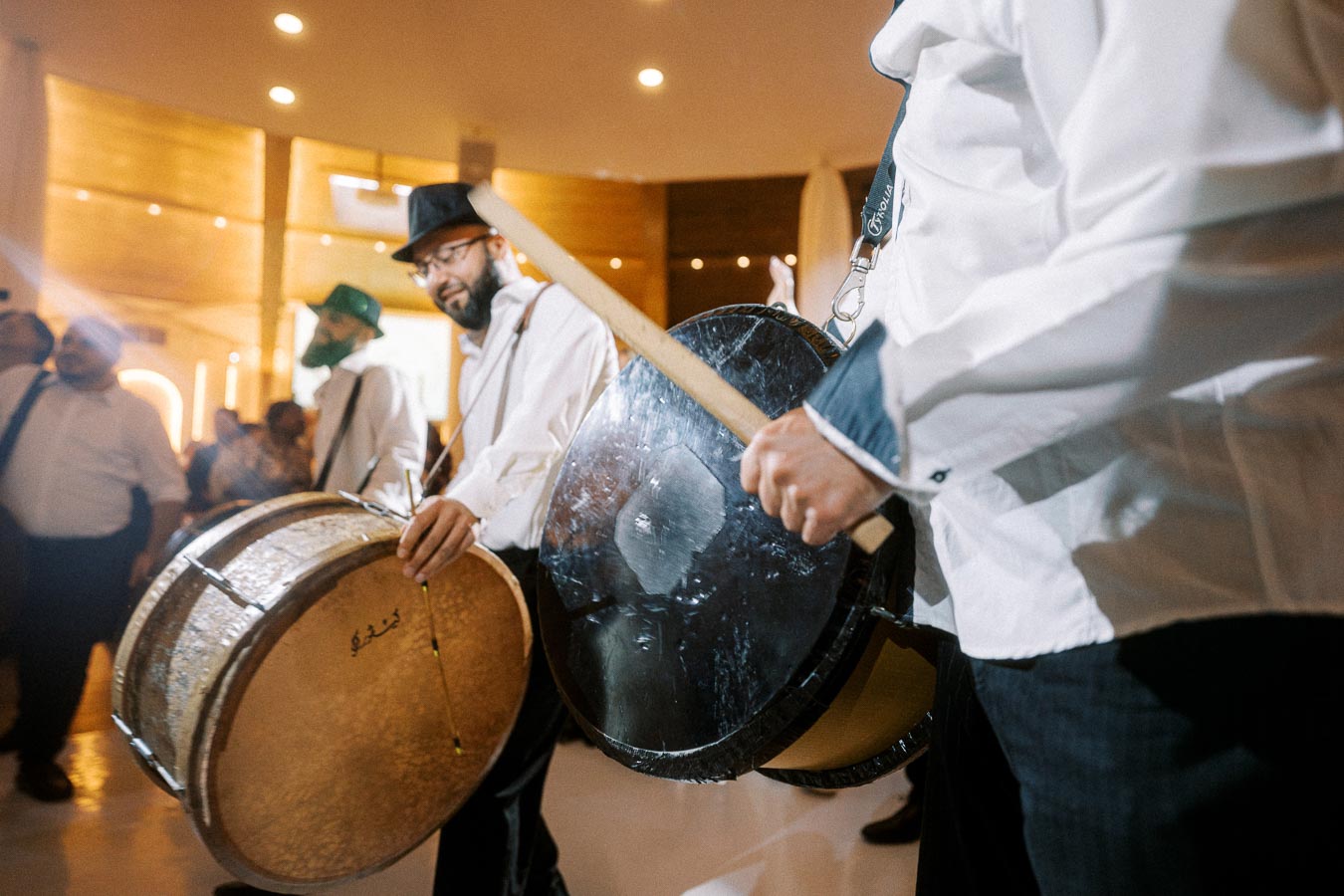Musicians energetically playing traditional drums at an indoor event, wearing white shirts and hats, creating a lively