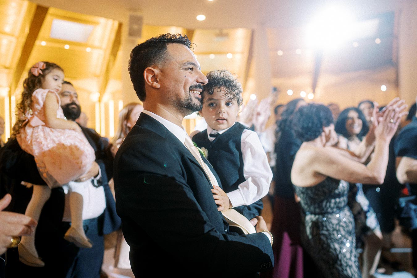 Man in a suit holding a young boy in formal attire at a celebration event, surrounded by guests clapping and smiling, in a