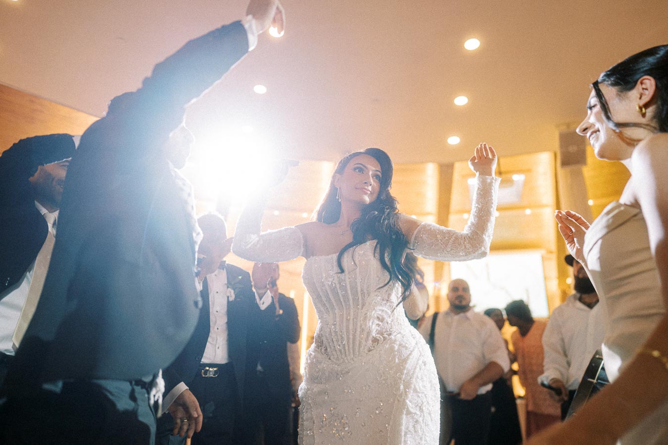Bride joyfully dancing with guests at a wedding reception, wearing a detailed white gown, surrounded by people