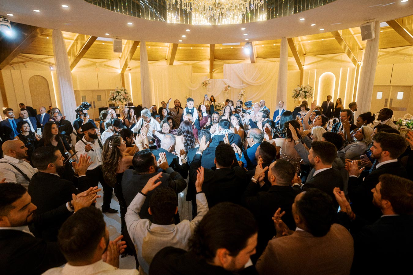 A large group of people celebrating at a wedding reception inside a beautifully decorated ballroom with elegant lighting and
