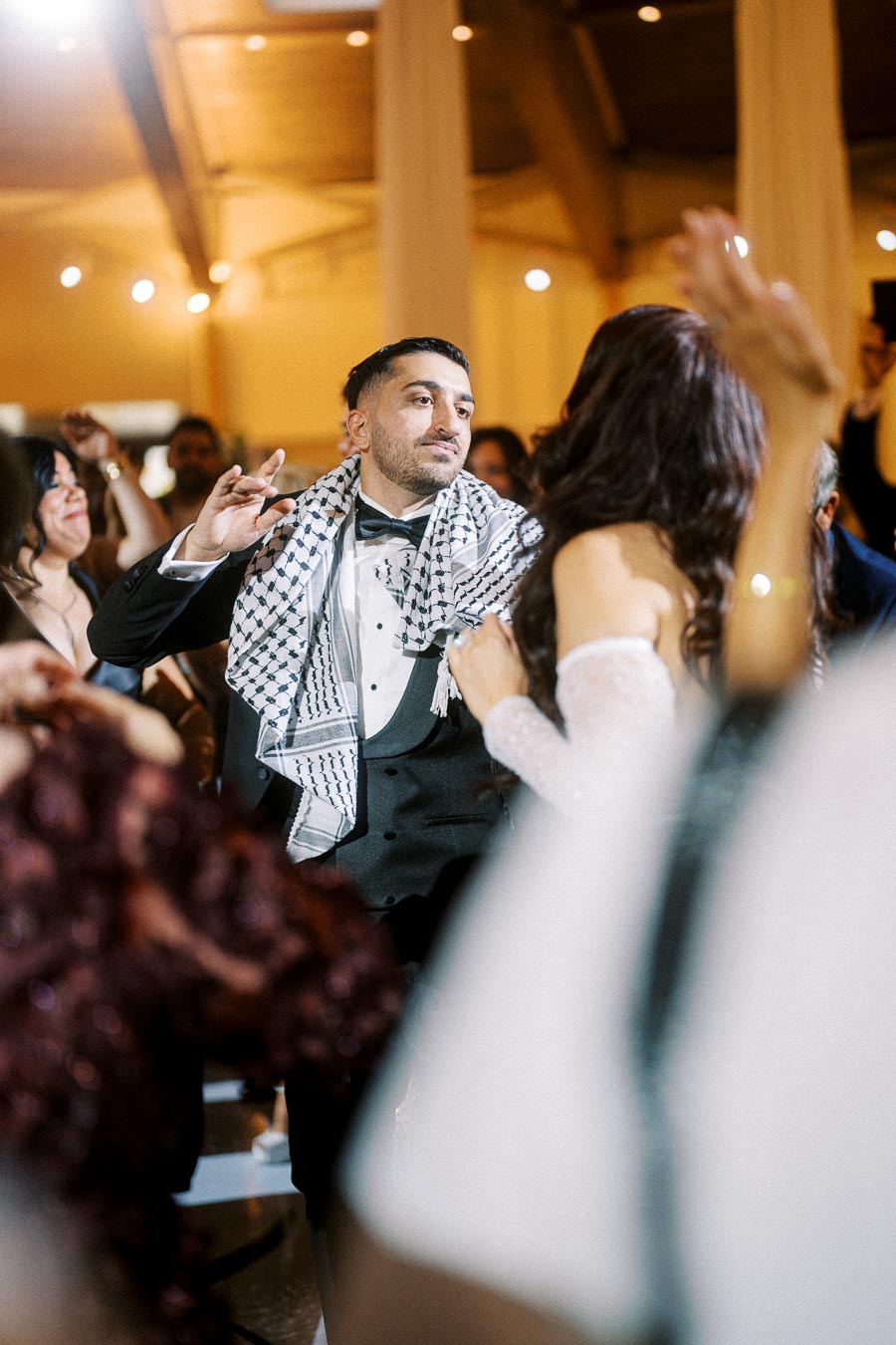 A man in a tuxedo with a keffiyeh draped over his shoulders joyfully dancing at a lively wedding reception, surrounded by