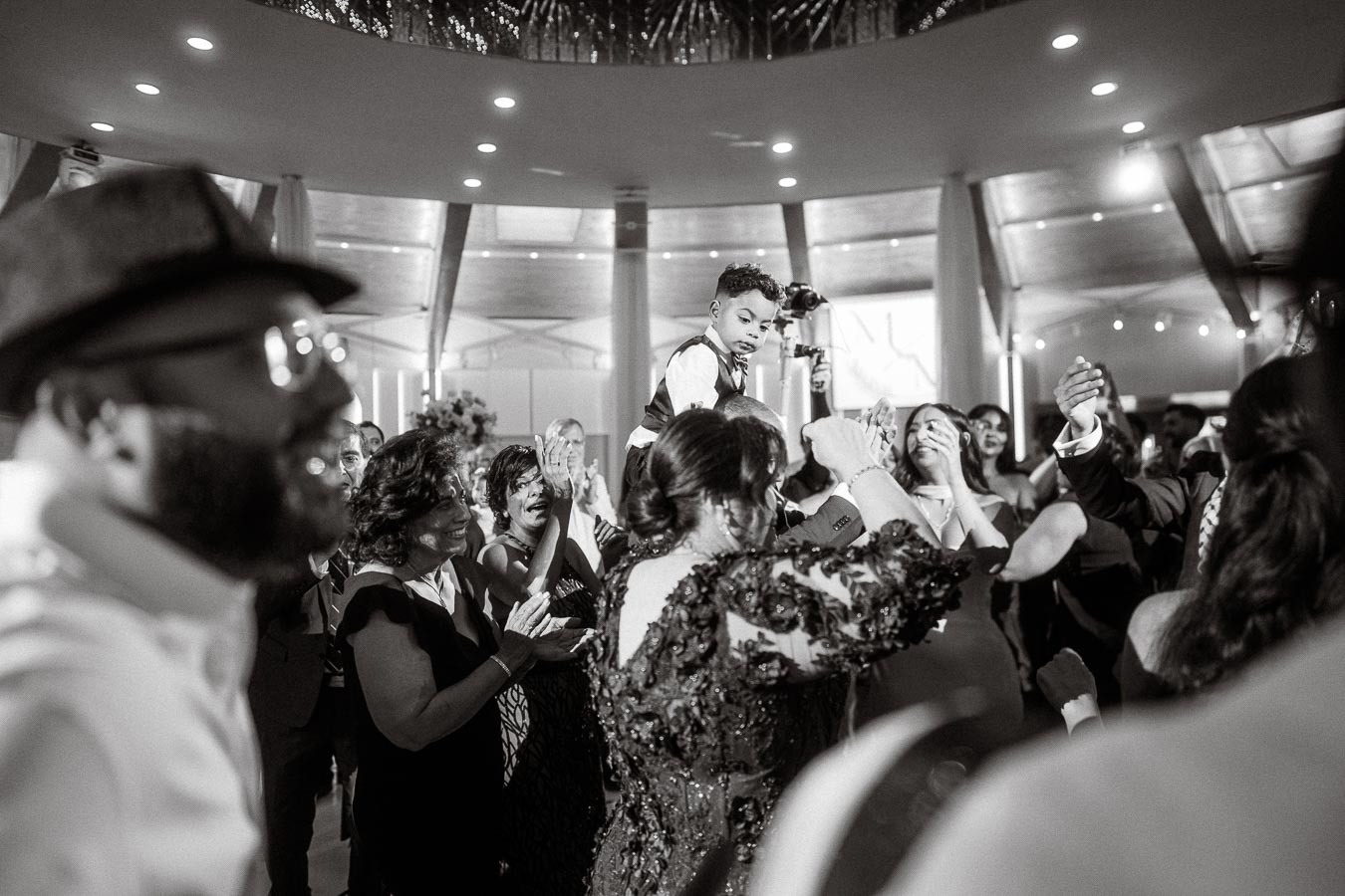 Black and white photo of a joyous celebration at a wedding reception, featuring a well-dressed young boy being lifted above