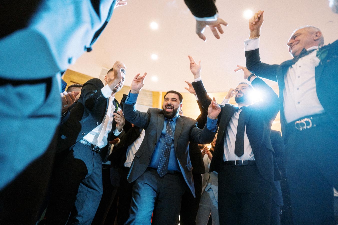Group of men in formal suits joyfully dancing at an indoor event reception.