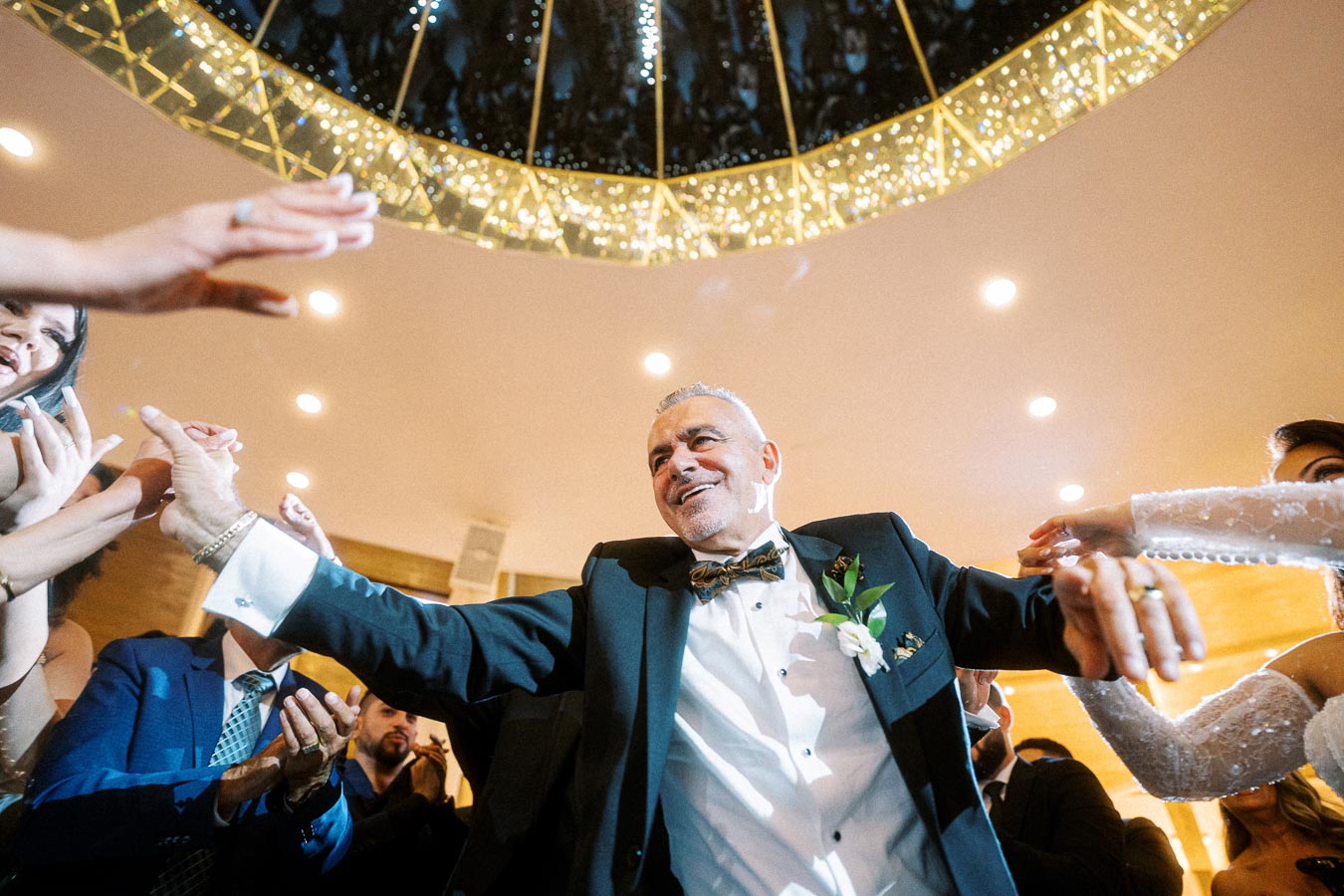 A joyful man in a formal suit and bowtie is dancing energetically at a wedding reception, surrounded by smiling guests under