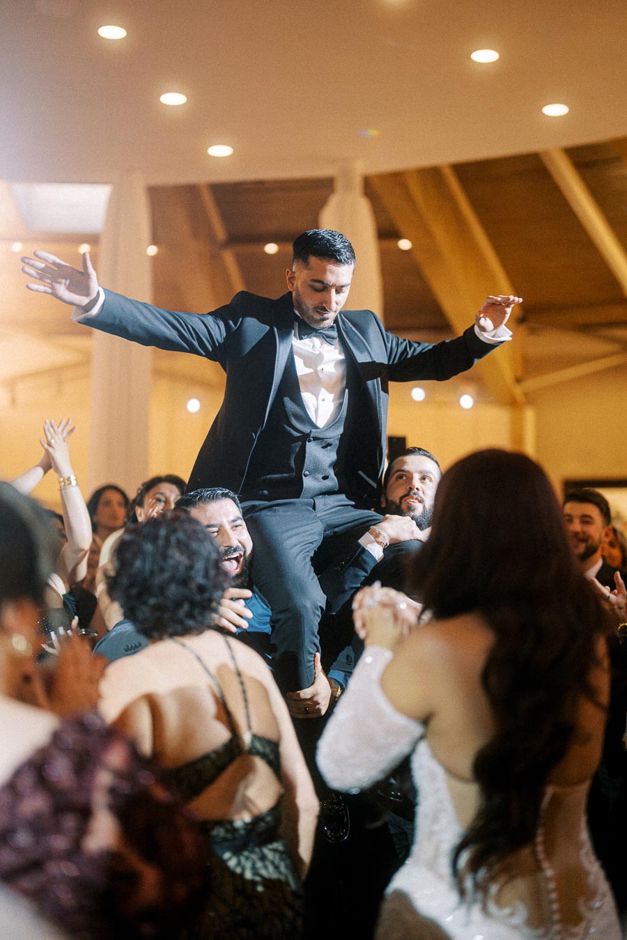 Man in a tuxedo being lifted by a joyful crowd at a lively wedding reception, surrounded by dancing guests and elegant decor.