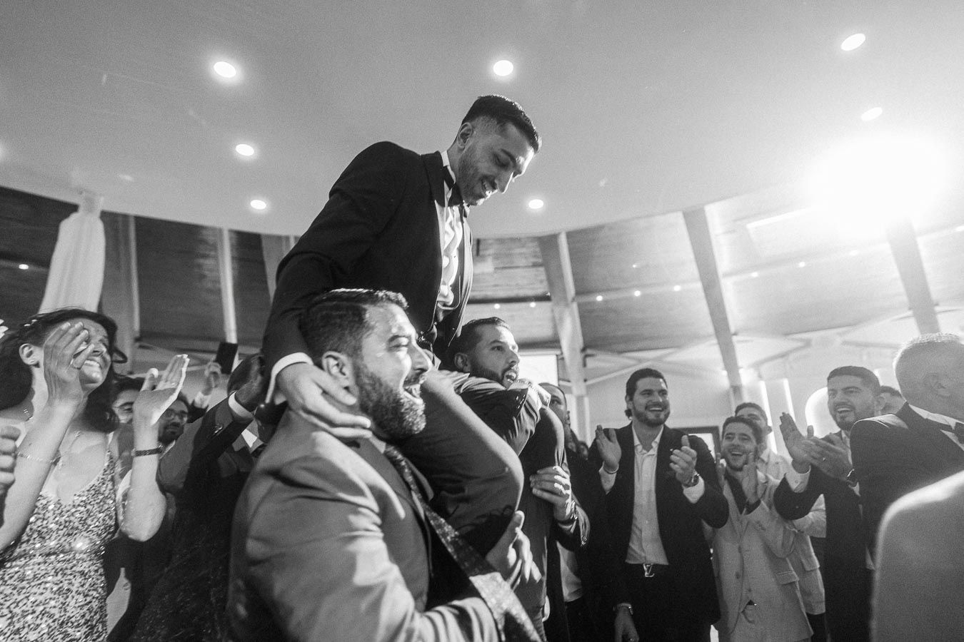 A group of people celebrating at a wedding, with a man in a suit being lifted by others in a joyful and lively atmosphere.