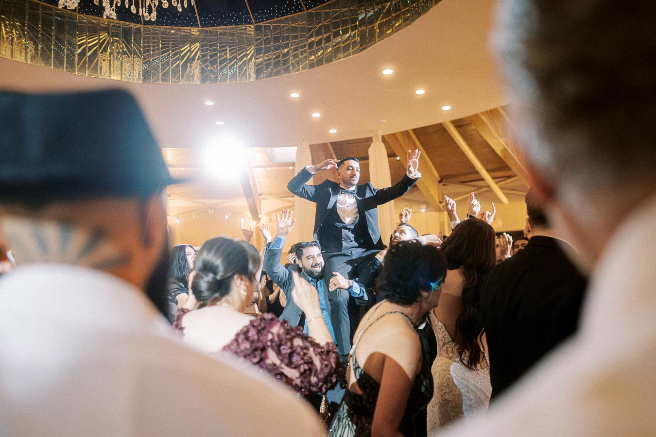 A joyous wedding celebration with the groom in a black tuxedo lifted on guests' shoulders, surrounded by dancing friends and