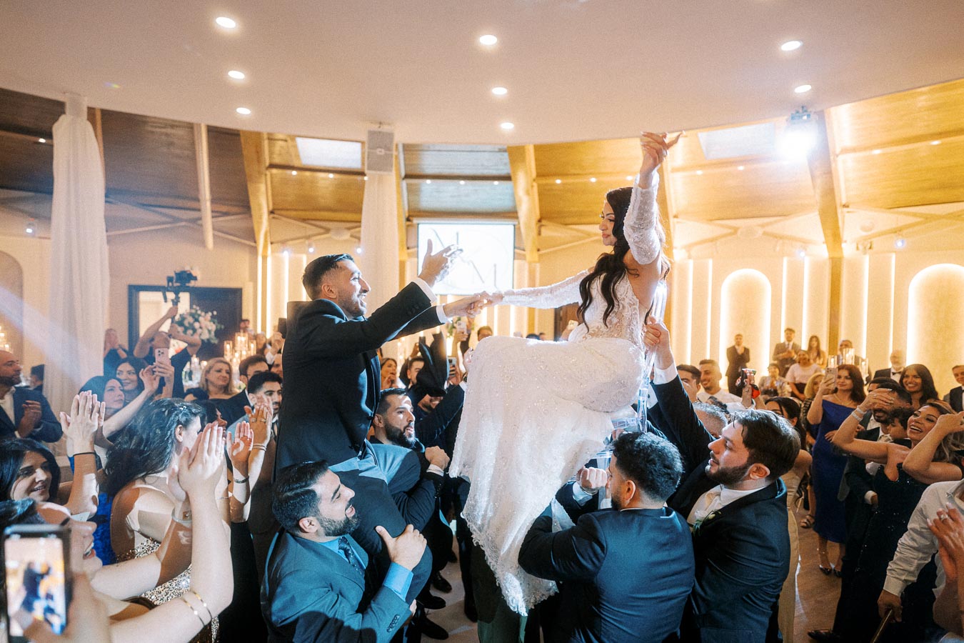 A joyful wedding celebration with a bride and groom being lifted up on chairs by guests, surrounded by a cheering crowd in a