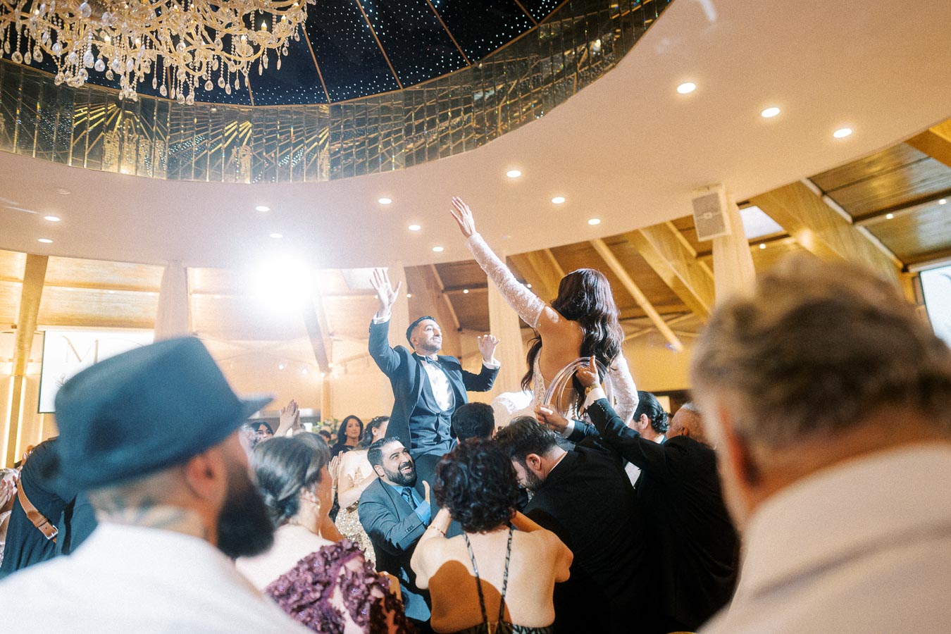 Group of people celebrating at a wedding reception, with a bride and groom being lifted on chairs under a sparkling