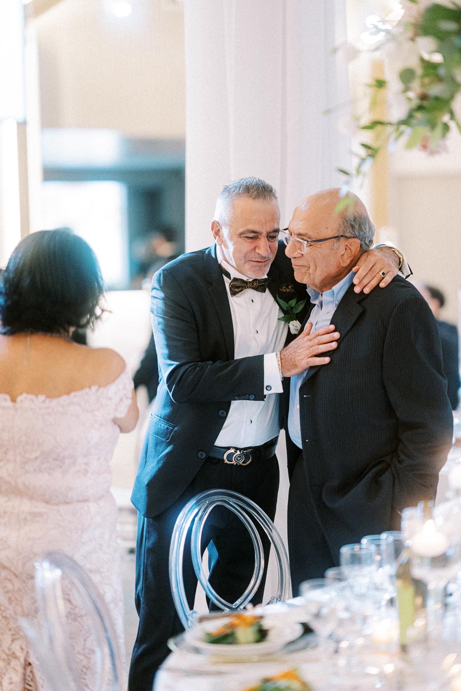 An elderly man in a tuxedo embraces another elderly man in a suit at a formal event, with a woman in a pink lace dress