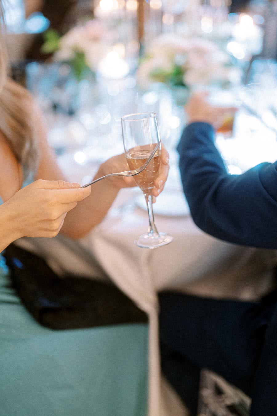 A close-up of two people at a formal event, one holding a champagne glass with a spoon ready to tap for attention,