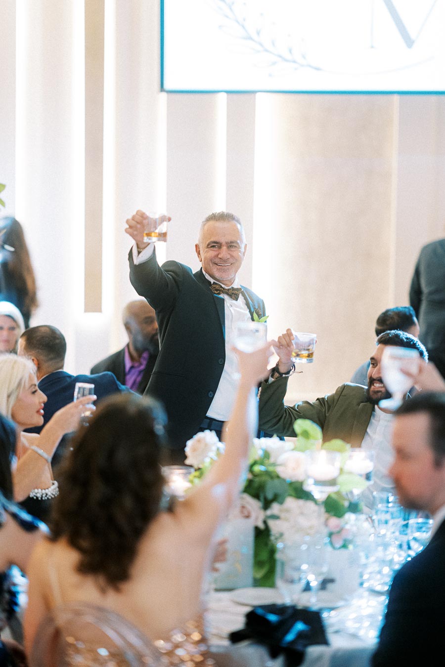 A wedding guest wearing a suit raises a glass during a toast at a beautifully decorated reception, surrounded by people at