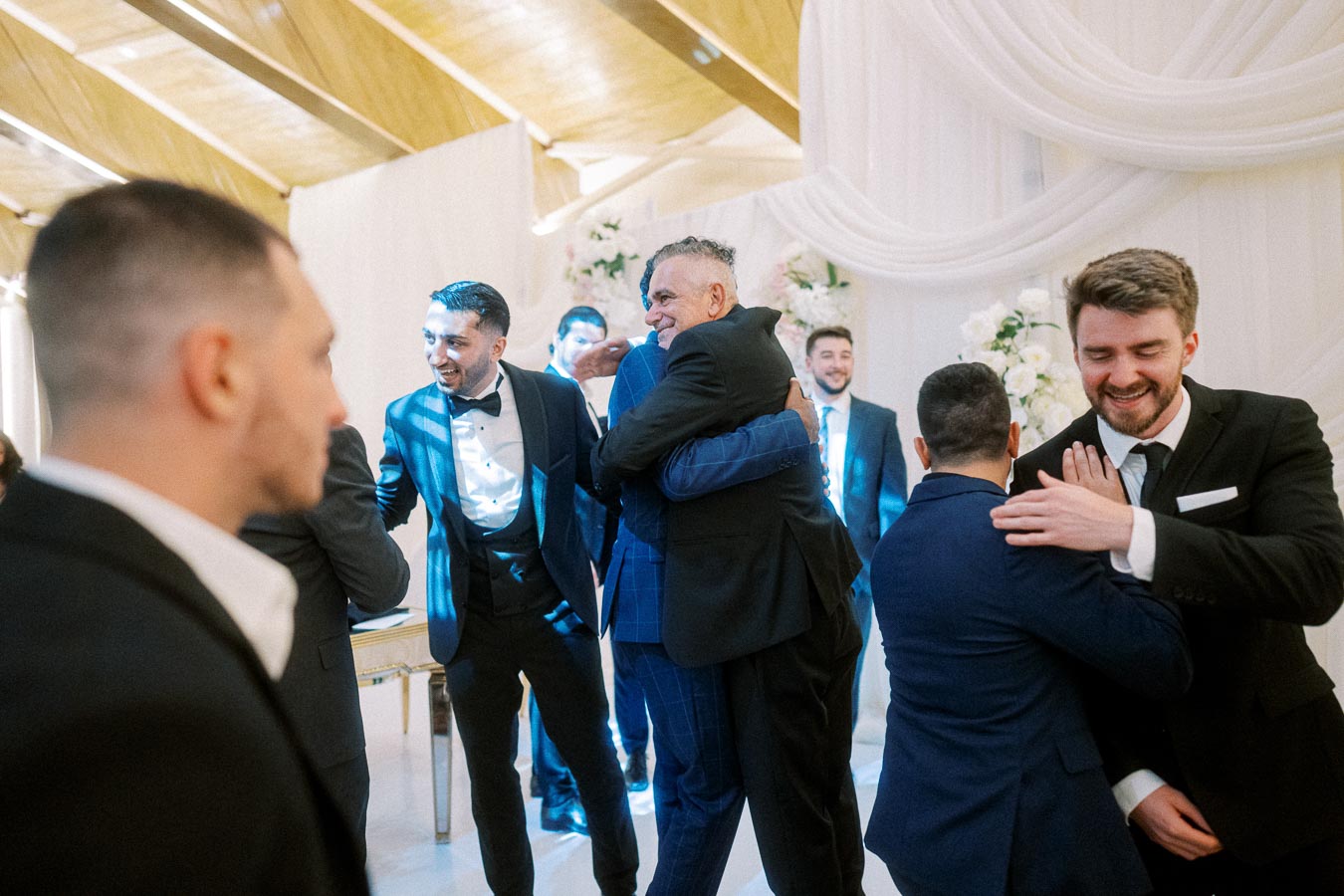 Group of men in suits hugging and congratulating each other at an elegant indoor event, with white draped decor and flowers