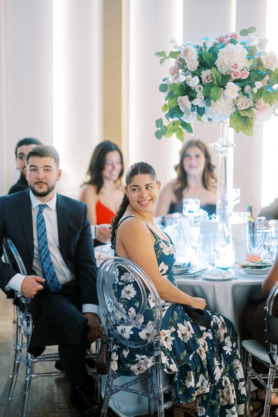 Guests seated at an elegant wedding reception, with a smiling woman in a floral dress and a man in a suit, surrounded by