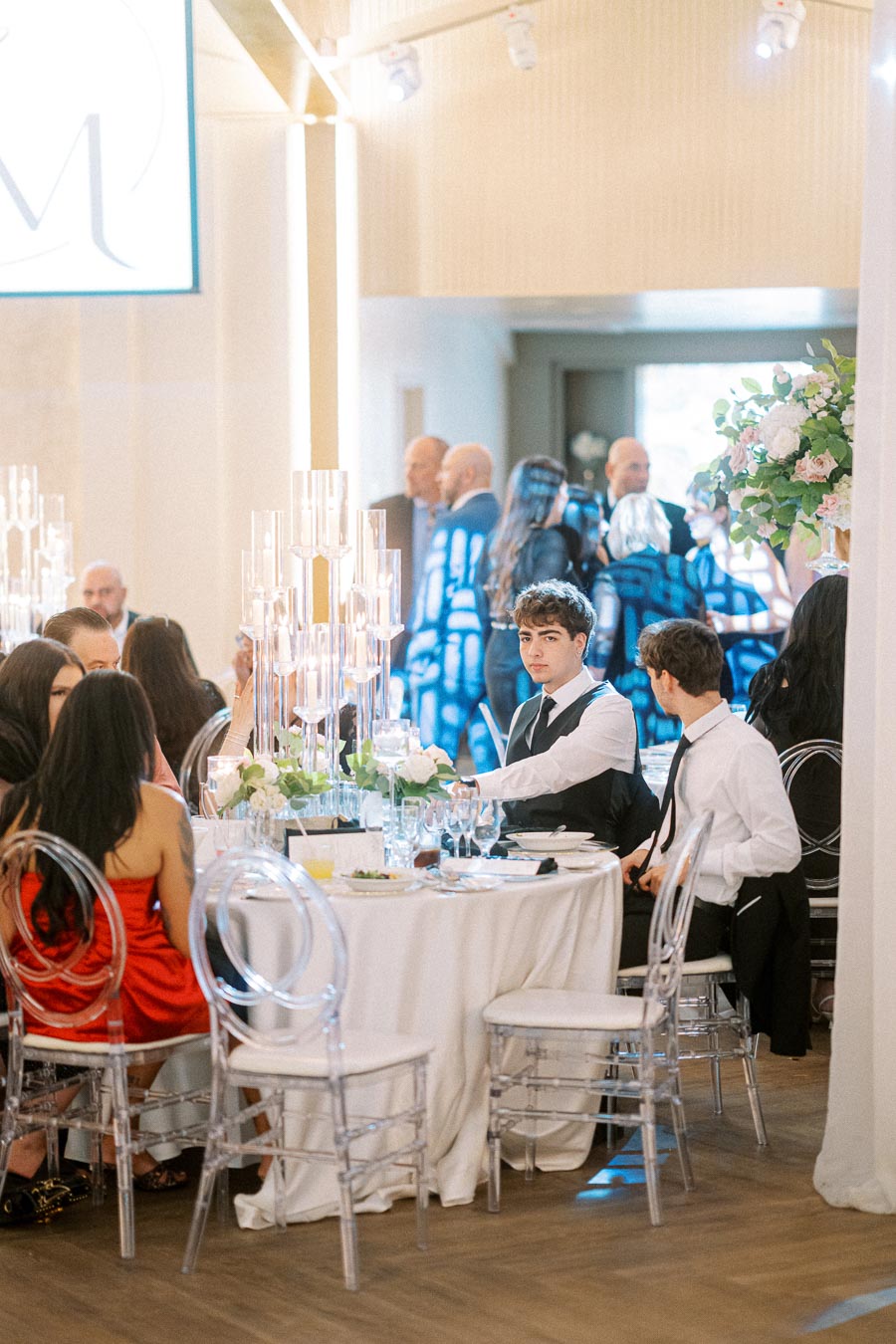 Elegant wedding reception with guests seated at tables adorned with white tablecloths, clear chairs, and floral
