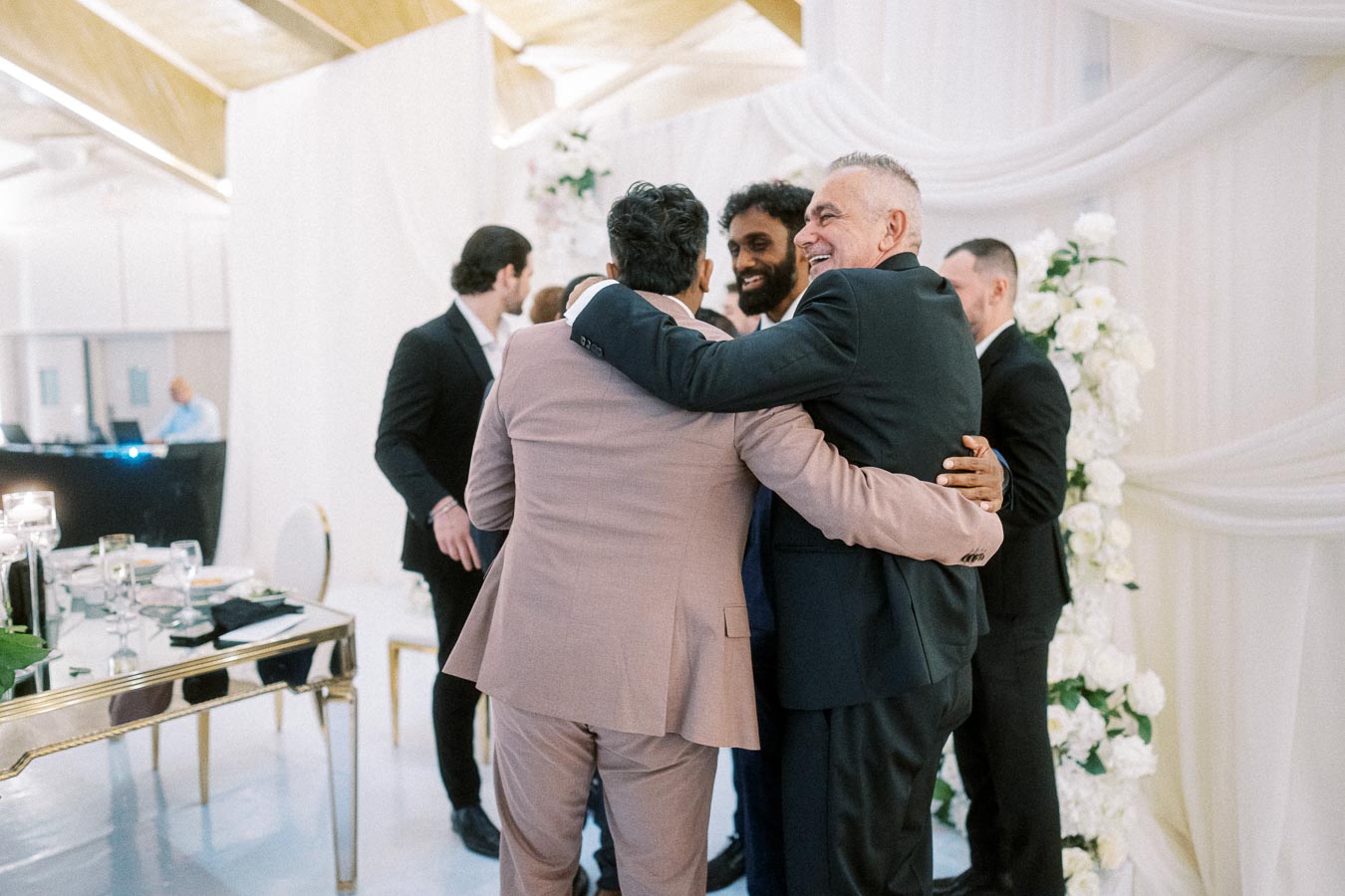 A group of well-dressed men embracing at a formal event, set in an elegantly decorated venue with draped fabric and floral