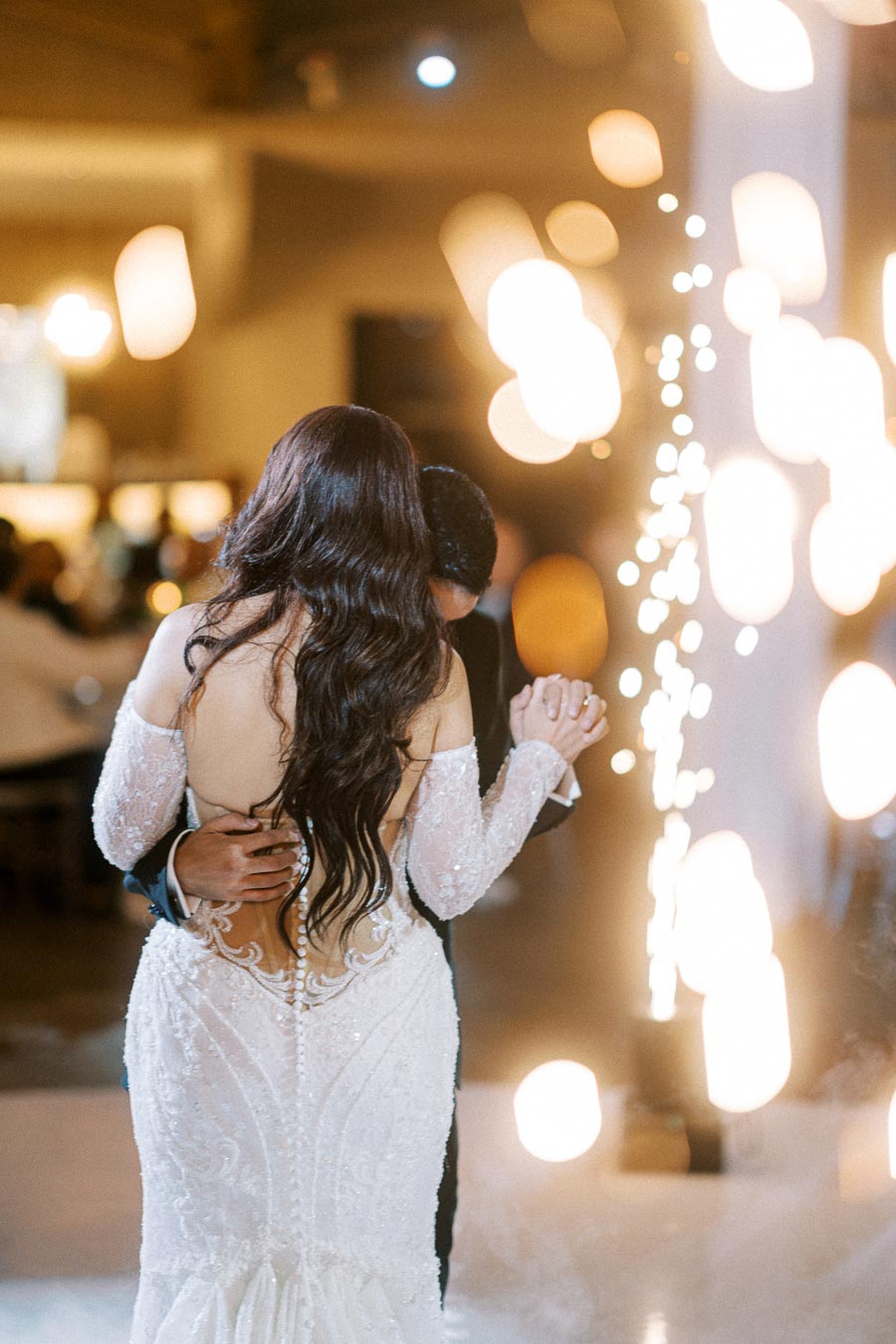 A bride in a stunning white gown shares a romantic first dance with her groom at a wedding reception, surrounded by blurred