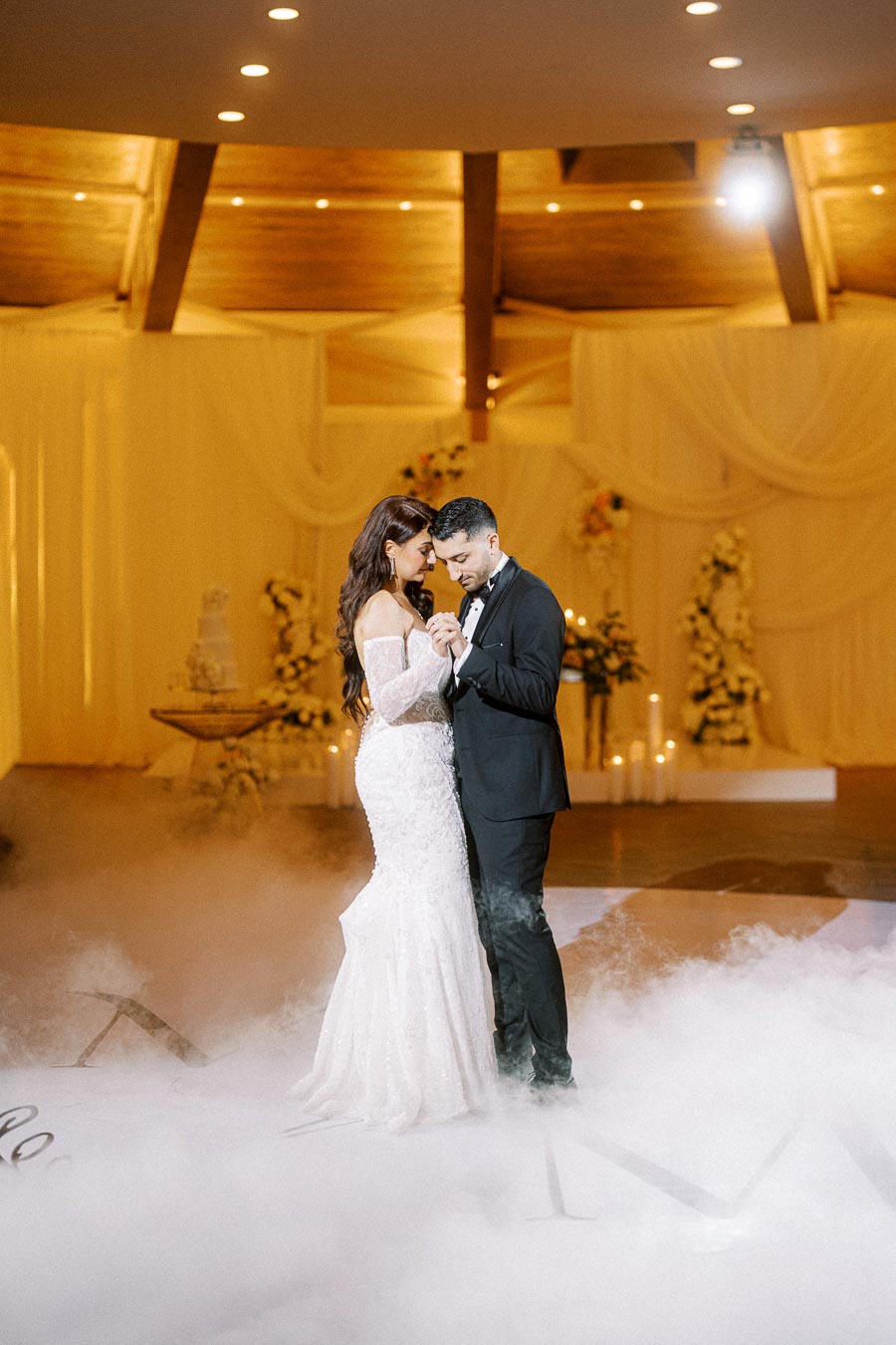 A bride and groom sharing a romantic first dance on the wedding floor, surrounded by soft ambient lighting and fog, in an