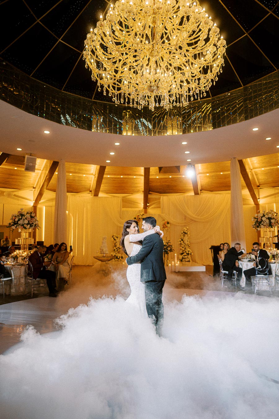 A couple shares their first dance under a grand chandelier in an elegantly decorated wedding venue, surrounded by mist and