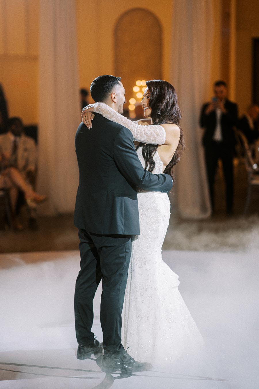 A bride and groom sharing their first dance in an elegant ballroom, surrounded by soft lighting and gentle smoke, with