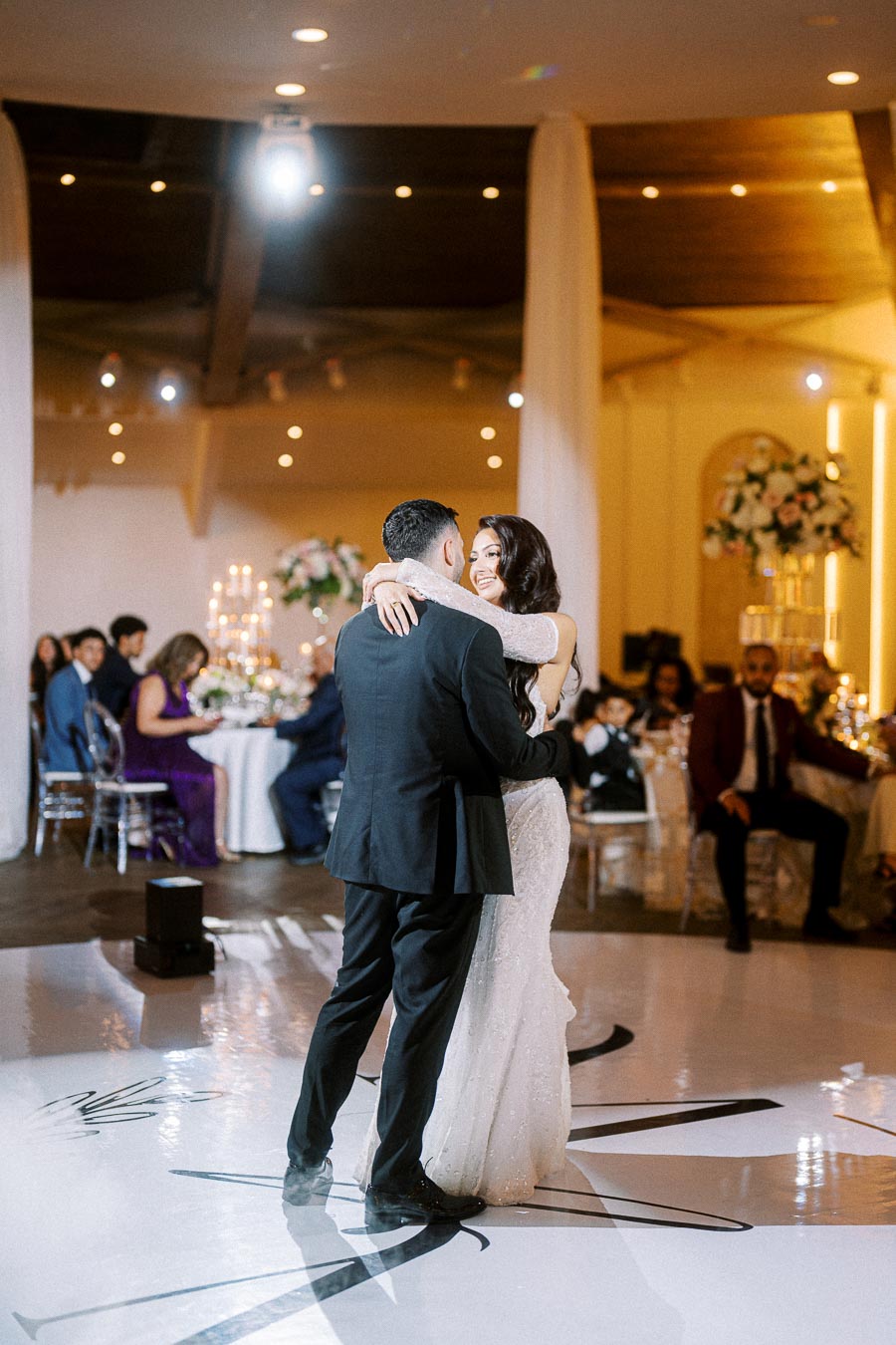 A bride and groom share a romantic first dance at their wedding reception, surrounded by elegantly decorated tables and