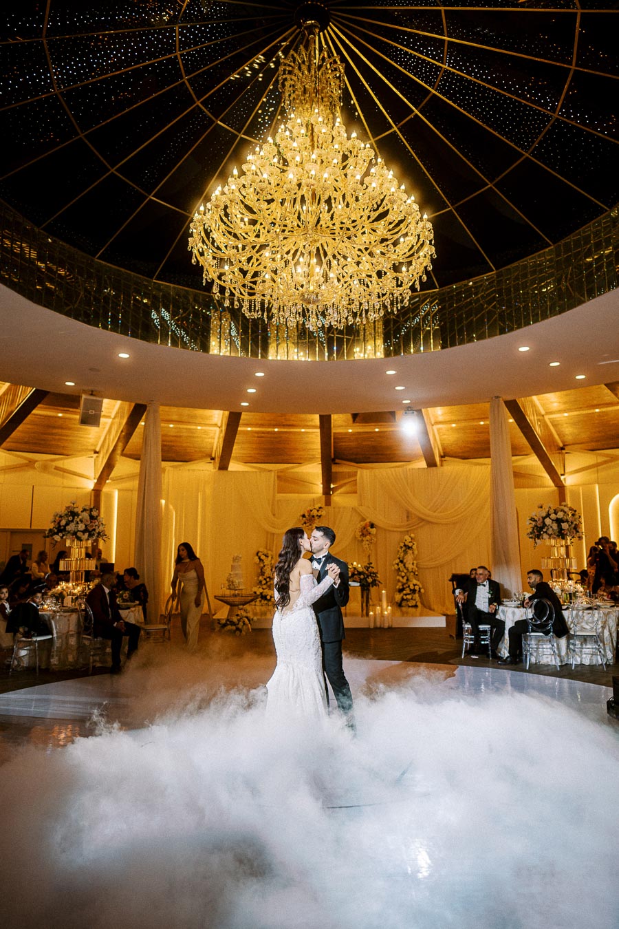 Elegant wedding reception with a couple sharing their first dance under a grand chandelier, surrounded by fog and guests in