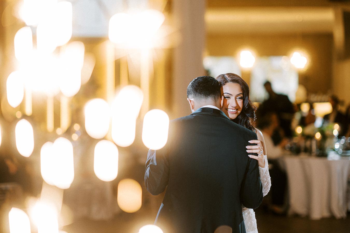 A couple shares their first dance at a wedding reception, surrounded by warm, ambient lighting; the bride smiles brightly