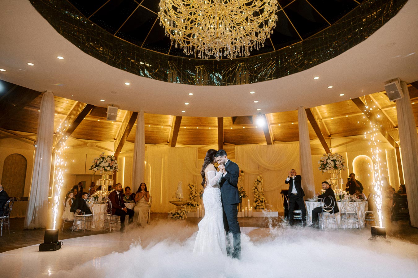 A bride and groom share their first dance under a magnificent chandelier in an elegantly decorated venue, with guests seated