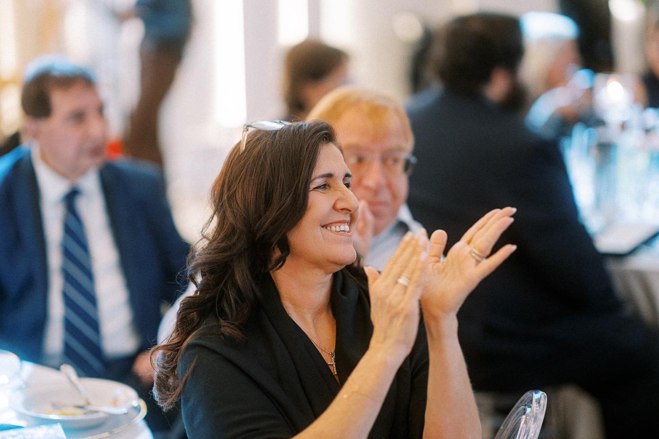 Woman smiling and clapping at a formal event, surrounded by seated attendees in business attire.