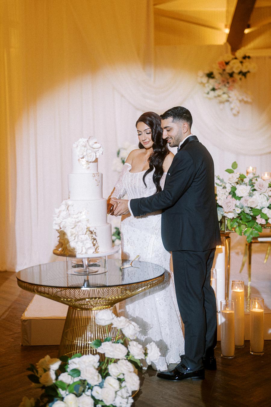 Couple cutting a tiered white wedding cake adorned with floral decorations, surrounded by elegant flower arrangements and