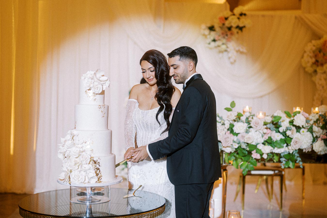 A couple in elegant wedding attire cuts a tiered white cake adorned with floral decorations, set against a backdrop of white