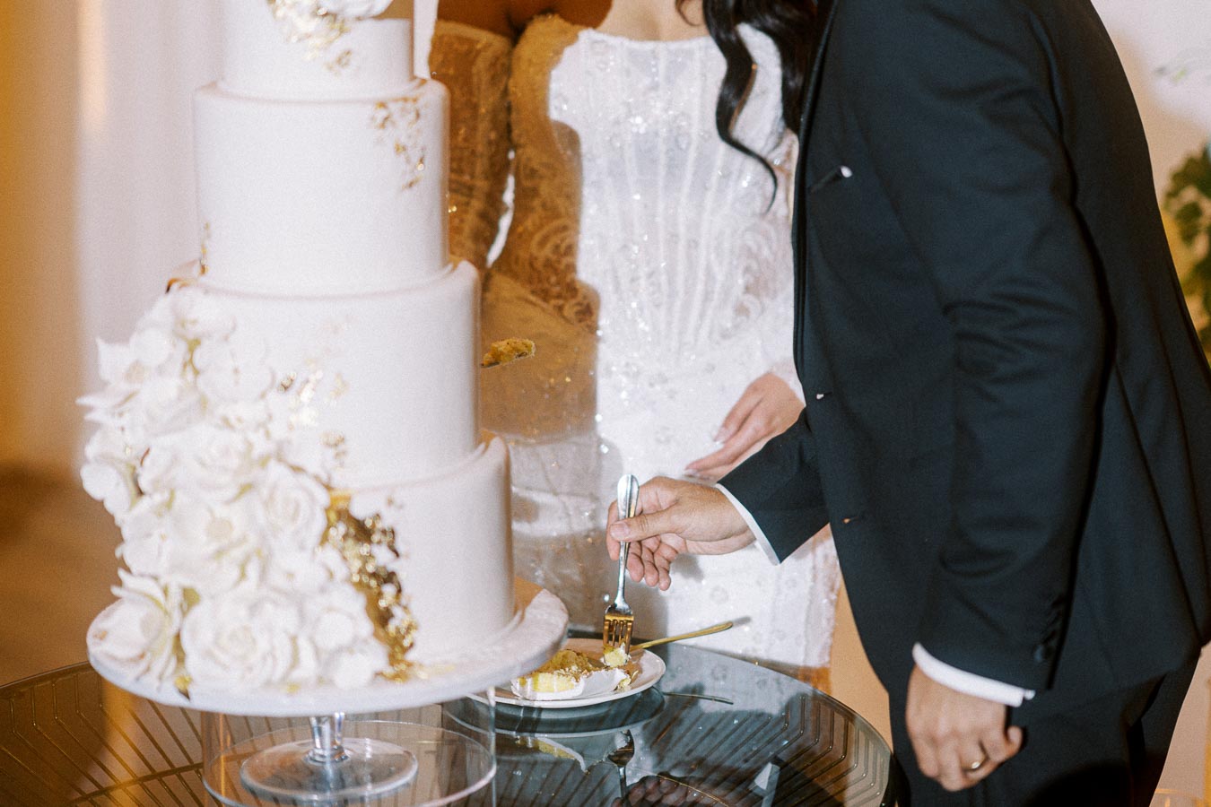 A bride and groom cutting a white, multi-tiered wedding cake adorned with gold accents and white flowers. The groom is
