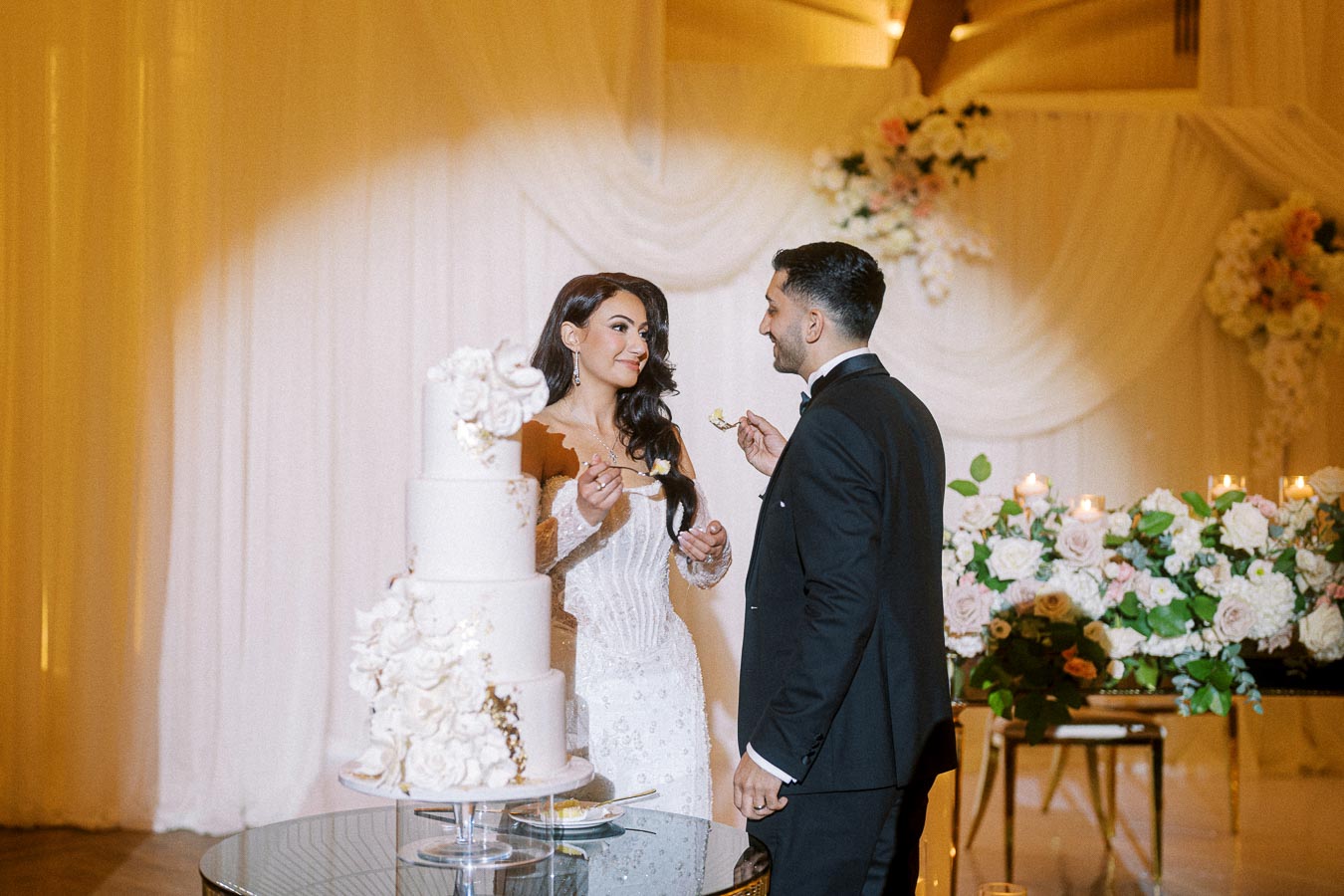 A bride and groom share a moment while cutting their elegant white wedding cake, adorned with floral decorations, at a